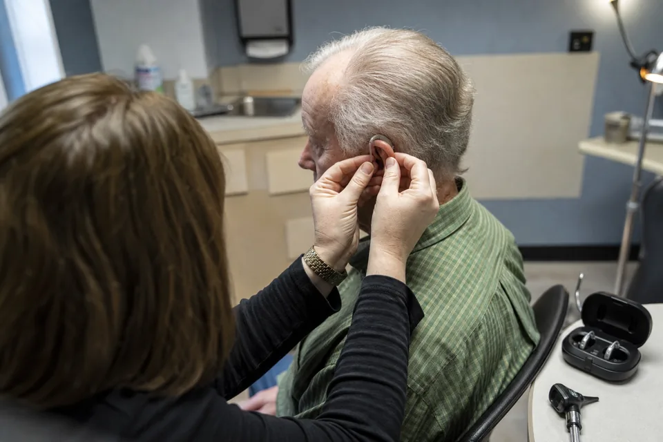 Audiologist fitting a hearing aid behind the ear of an older patient in a clinic exam room